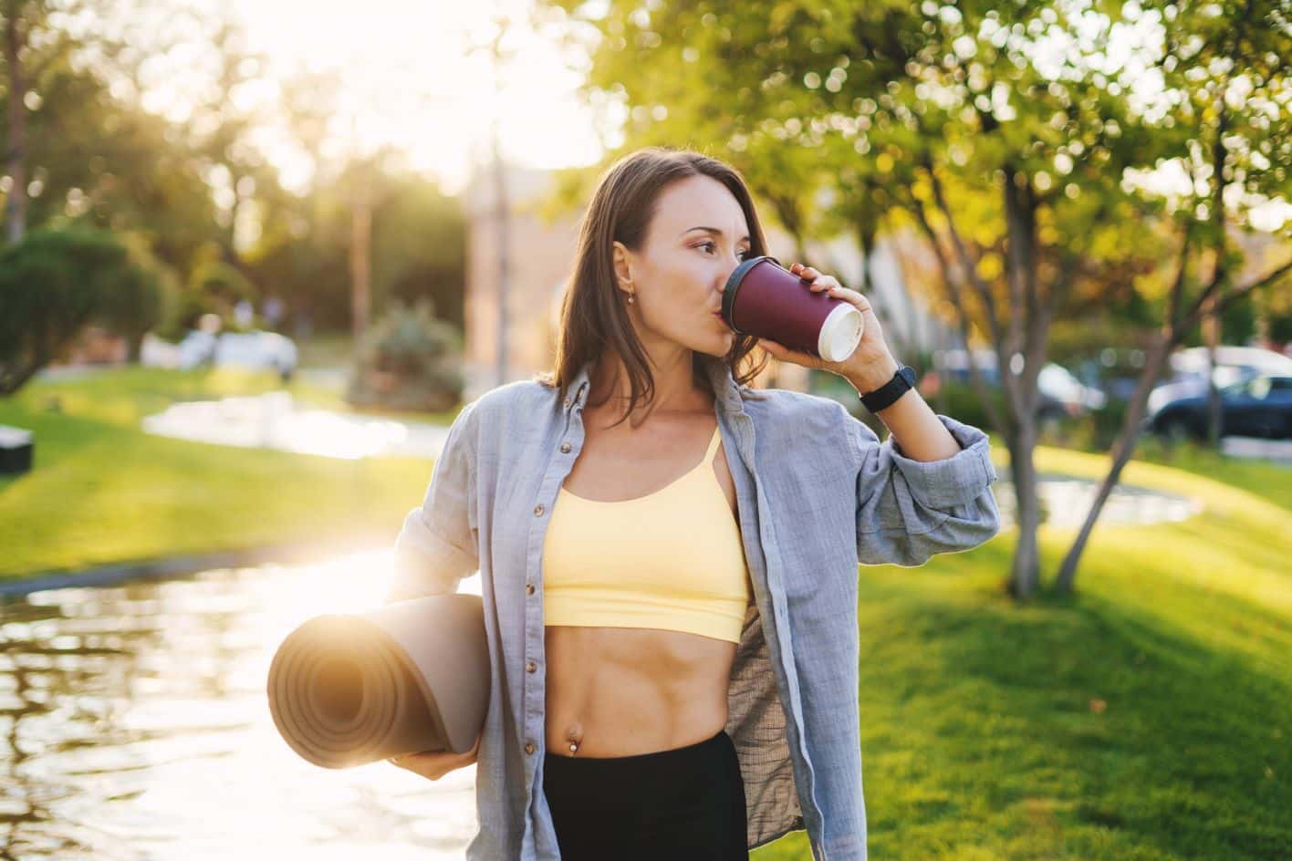 A woman wearing a yellow sports bra and open button-down shirt is standing outdoors with a yoga mat under her arm, drinking from a disposable cup. She is in a park-like setting with green grass, trees, and a pond in the background. MyFitnessPal Blog